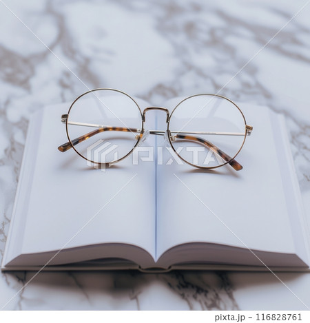 A pair of round eyeglasses rests on an open book, the pages are blank and the background is a minimalist marble surface. 116828761