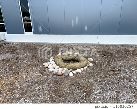 This image depicts an outdoor drainage system protected by erosion control measures. The drain is surrounded by a circular barrier made of natural fiber rolls and large river rocks. The setup ensures 116830257