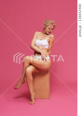 Blonde woman wearing white lingerie posing sitting on wooden block and touching her legs against pink background. Epilation. 116832488