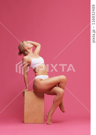 Contemplative woman in minimal white lingerie is seated on wooden block, radiating elegance against pink background. 116832489