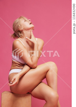 Serene blonde woman poses in white lingerie, sitting on wooden block against pink studio background. 116832490