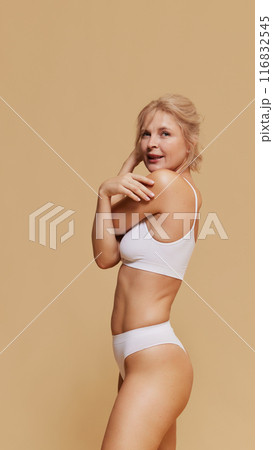 Blonde woman in white underwear poses gracefully, her hand on her shoulder against pastel studio background. Blonde woman in white underwear poses gracefully, her hand on her shoulder against pastel studio background. 116832545