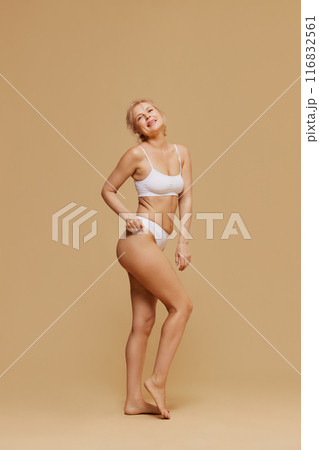Joyfully woman in white underwear strikes playful pose with her hand on her hip against pastel studio background. 116832561