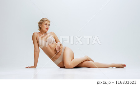 Elegant woman in beige lingerie lying, posing on floor against white studio background, exuding beauty an femininity. 116832623