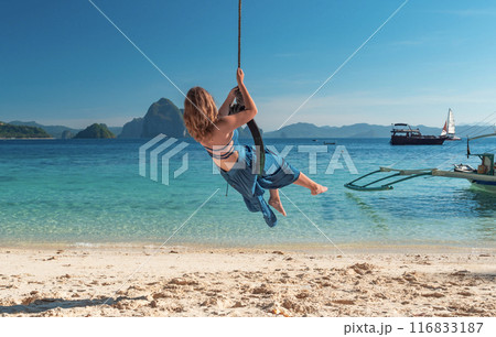 A woman with long blonde hair, wearing a blue dress, swings on a tire swing over crystal clear turquoise waters. 116833187