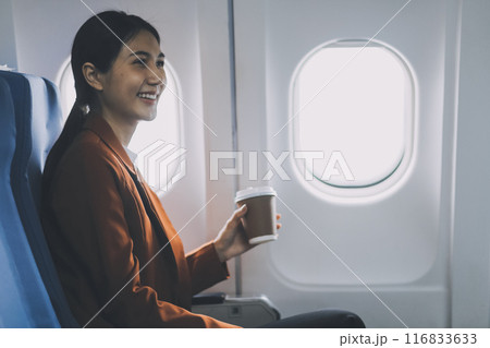 Smiling Asian woman enjoying smartphone comfortable flight while sitting in the airplane cabin, Passengers near the window. 116833633