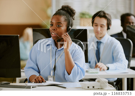African American female call center operator communication with client via headphones while coworkers working in blurred background African American female call center operator communication with client via headphones while coworkers working in blurred background 116833753