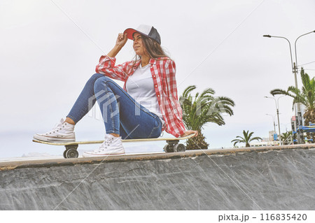 Beautiful young woman in cap is sitting at skatepark on the ramp with her longboard. Beautiful young woman in cap is sitting at skatepark on the ramp with her longboard. 116835420