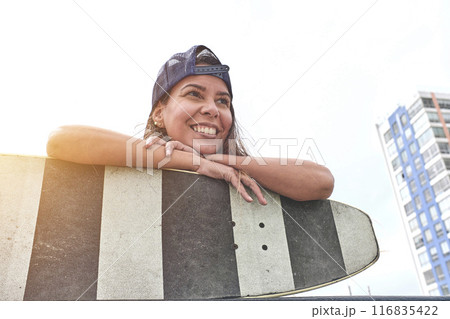 Beautiful young woman in cap is sitting at skatepark on the ramp with her longboard. Beautiful young woman in cap is sitting at skatepark on the ramp with her longboard. 116835422