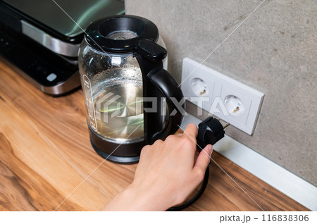 glass modern electric kettle stands on the table against the background of an outlet. Boiling water. Electricity consumption by household electrical appliances glass modern electric kettle stands on the table against the background of an outlet. Boiling water. Electricity consumption by household electrical appliances 116838306
