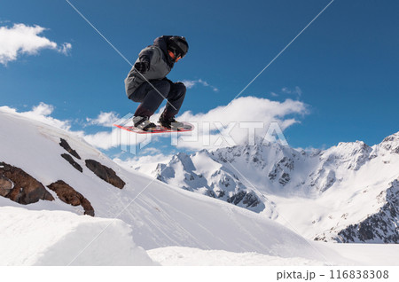 Jumping snowboarder holds one hand on a snowboard against the background of a blue sky and large snow-capped mountains. Performing a stunt in flight Jumping snowboarder holds one hand on a snowboard against the background of a blue sky and large snow-capped mountains. Performing a stunt in flight 116838308