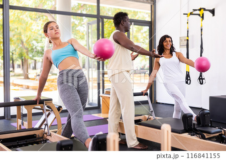 Attentive African American male trainer helping two positive women training with pilates bender ball Attentive African American male trainer helping two positive women training with pilates bender ball 116841155
