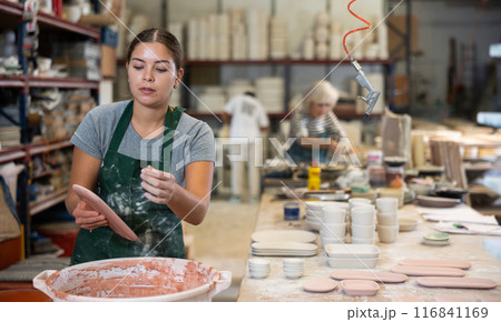 Young female worker in pottery factory lowers fired clay plates and cups into container with glaze Young female worker in pottery factory lowers fired clay plates and cups into container with glaze 116841169