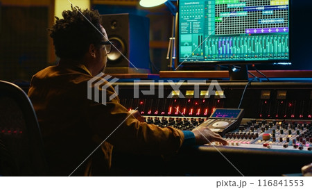African american sound expert operates on audio console with moving faders and colored meters, editing in professional studio control room. Producer works on album, recording hit song. Camera A. African american sound expert operates on audio console with moving faders and colored meters, editing in professional studio control room. Producer works on album, recording hit song. Camera A. 116841553