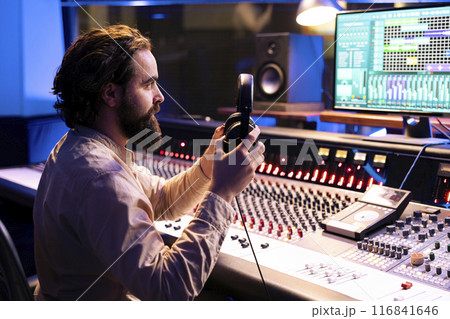 Audio technician putting headphones on to listen to final track in studio, editing records with faders an knobs on control panel desk. Professional music producer creating new songs, post production. Audio technician putting headphones on to listen to final track in studio, editing records with faders an knobs on control panel desk. Professional music producer creating new songs, post production. 116841646