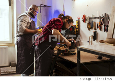 Master carpenter in joinery watching apprentice using orbital sander on lumber. Team in woodworking shop using angle grinder to create wooden decorations, refining wood objects 116841709