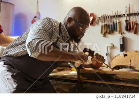 BIPOC man in carpentry shop wearing protection equipment while carving wood to prevent workplace accidents. Cabinetmaker equipped with safety goggles while using chisel and hammer to avoid injury 116841714