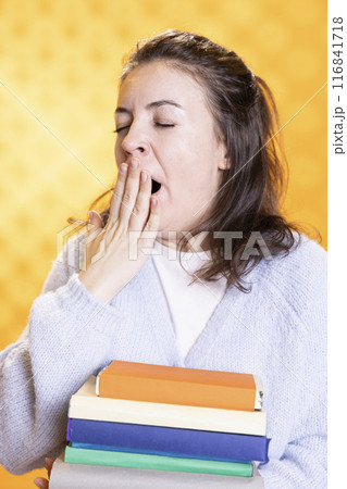 Woman yawning, feeling exhausted from stress, carrying books for school exam, studio background. Tired student feeling sleepy after doing homework all night before university assessment 116841718