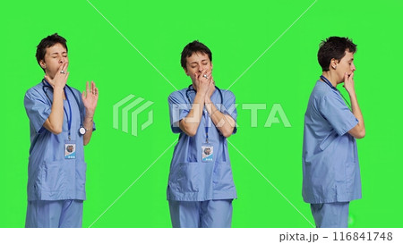 Medical assistant feeling extremely sleepy against greenscreen backdrop, being overworked after the night shift. Woman nurse yawns and feels exhausted in studio, burnout and anxiety. Camera B. 116841748