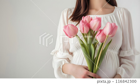 A pregnant woman in a white dress holding a bouquet of pink tulips celebrates motherhood and new life, embracing elegance and femininity in a delicate and floral setting 116842300