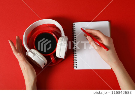 A cup of coffee with headphones and a notebook on a red background 116843688