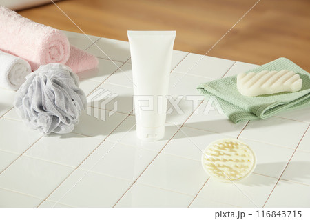 Bath cosmetic advertising photo with high close up shot, a white blank label tube displayed on a checkered table top of brick, decorated by some bathroom items like towel, soap, bath sponge and brush 116843715