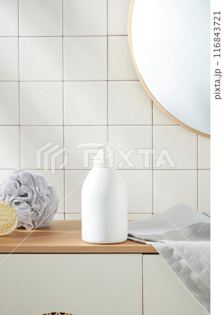 Close up shot photography at an unlabeled shower gel bottle in white color, which placed above of wooden cabinet. On both side, displaying some bathroom accessories such as towel and bath sponge 116843721