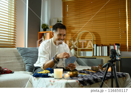 Smiling millennial man conducting a tarot reading session while live streaming in living room Smiling millennial man conducting a tarot reading session while live streaming in living room 116850117
