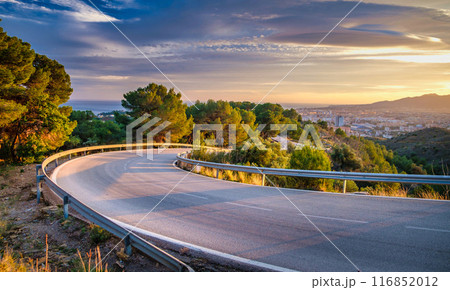 winding road that curves gently to the left as it meanders through a scenic landscape. The road is bordered by guardrails for safety and is surrounded by lush greenery 116852012