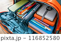 Professional photograph of a school backpack brimming with supplies. The backpack, positioned upright, is slightly open, showcasing various school essentials. Visible items include spiral notebooks, 116852580