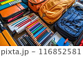 Professional photograph of a school backpack brimming with supplies. The backpack, positioned upright, is slightly open, showcasing various school essentials. Visible items include spiral notebooks, 116852581