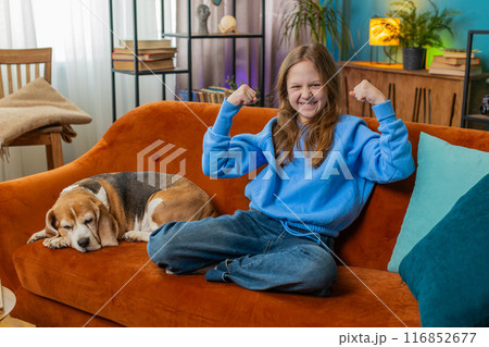 Cheerful young child girl celebrating victory winning the lottery at home beside beagle dog on sofa 116852677