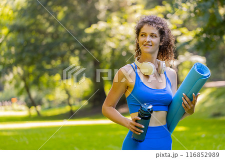 Young happy sportswoman with mat and water bottle in hands is heading to training outdoors in park Young happy sportswoman with mat and water bottle in hands is heading to training outdoors in park 116852989