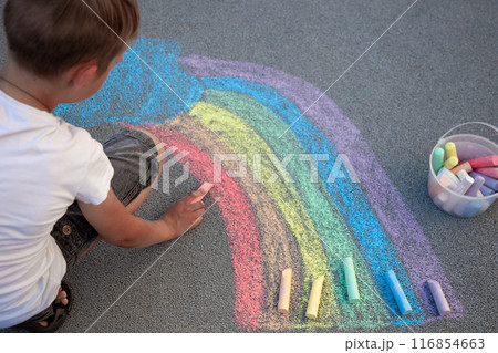 A boy draws a rainbow with multi-colored chalk on the asphalt. Drawing multicolored chalk stick, back to school background. Top view. 116854663