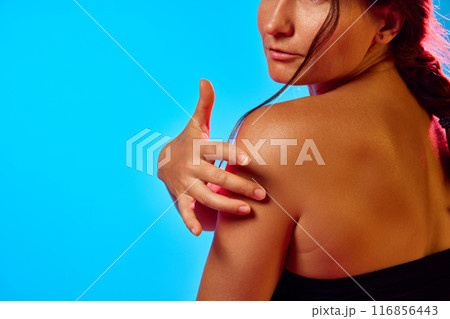 Back view of woman with braided hair, touching her shoulder, showcasing her smooth skin in neon light against blue background. 116856443