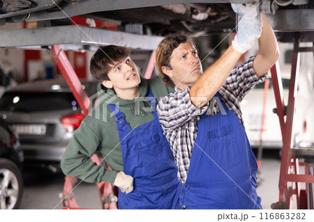 Young auto mechanic helps an experienced auto mechanic repair a car in car service center Young auto mechanic helps an experienced auto mechanic repair a car in car service center 116863282