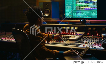 Tracking engineer processing and mixing sounds in control room with audio console and professional editing software on computer. Music production expert uses motorized faders and knobs. Camera A. Tracking engineer processing and mixing sounds in control room with audio console and professional editing software on computer. Music production expert uses motorized faders and knobs. Camera A. 116863887
