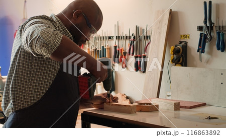 African american man in carpentry shop using power drill to create holes for dowels in wooden board. Craftsperson sinks screws into wood block with electric tool, doing precise drilling, camera A African american man in carpentry shop using power drill to create holes for dowels in wooden board. Craftsperson sinks screws into wood block with electric tool, doing precise drilling, camera A 116863892