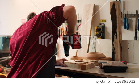 Woodworker in assembly shop using power drill to create holes for dowels in wooden board. Carpenter sinking screws into wooden surfaces with electric tool, doing precise drilling for seamless joinery Woodworker in assembly shop using power drill to create holes for dowels in wooden board. Carpenter sinking screws into wooden surfaces with electric tool, doing precise drilling for seamless joinery 116863912