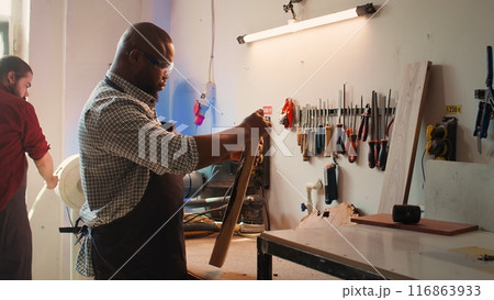 BIPOC carpenter in woodworking shop inspecting lumber piece before assembling furniture, checking for scratches. Cabinetmaker evaluates timber block, picking right tool from rack for it, camera B 116863933