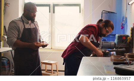 Carpenter making modifications on blueprint drawings, using angle grinder on wood. Man using orbital sander gear after consulting schematics, assisted by african american apprentice, camera B Carpenter making modifications on blueprint drawings, using angle grinder on wood. Man using orbital sander gear after consulting schematics, assisted by african american apprentice, camera B 116863957