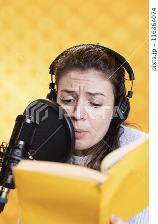 Woman frowns while doing voiceover reading of book to produce audiobook using melodramatic acting. Narrator glowers, using storytelling skills to portray character, recording novel, studio background Woman frowns while doing voiceover reading of book to produce audiobook using melodramatic acting. Narrator glowers, using storytelling skills to portray character, recording novel, studio background 116864074
