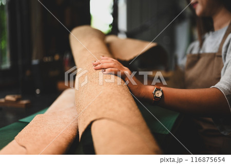 Hand of craftswoman examining a large roll of leather at her workbench surrounded by tools 116875654