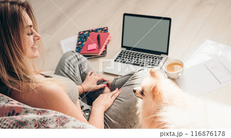 A young girl sits on the floor with her dog Pomeranian and works on a laptop. Dog is looking at her 116876178