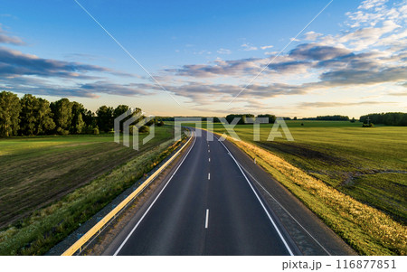 A winding road stretches into the distance, flanked by vibrant green fields and dense trees under a clear blue sky. A winding road stretches into the distance, flanked by vibrant green fields and dense trees under a clear blue sky. 116877851