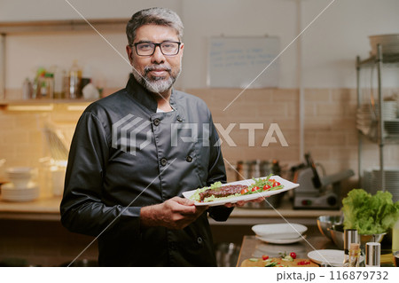 Proud middle aged chef dressed in black uniform smiling widely while demonstrating his served dish Proud middle aged chef dressed in black uniform smiling widely while demonstrating his served dish 116879732