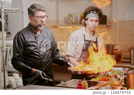 Middle aged chef cooking meat with fire while his best cook watching process Middle aged chef cooking meat with fire while his best cook watching process 116879738