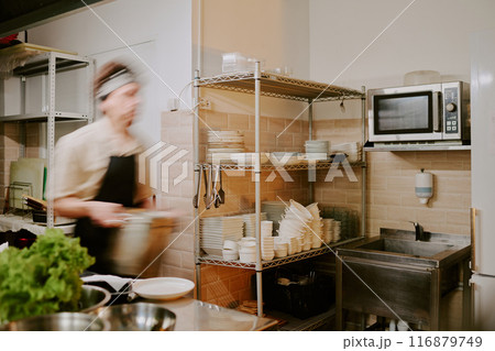 Young cook carrying huge steel pot in kitchen, kitchen fully equipped with cookware Young cook carrying huge steel pot in kitchen, kitchen fully equipped with cookware 116879749