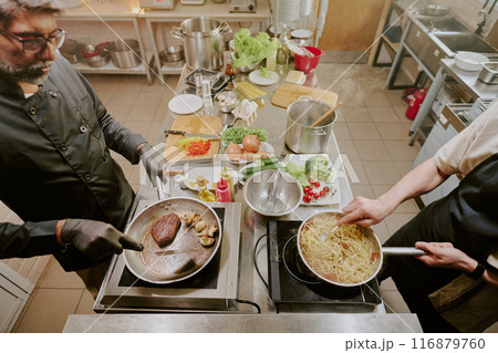 High angle shot of two professional cooks preparing italian pasta and black angus steak in modern kitchen High angle shot of two professional cooks preparing italian pasta and black angus steak in modern kitchen 116879760