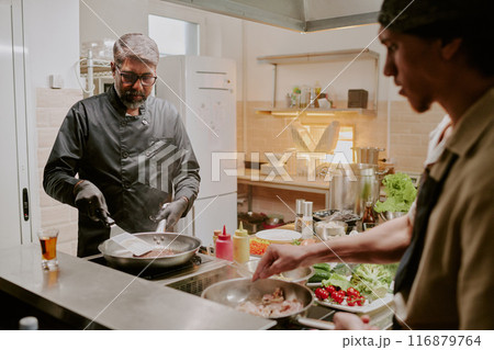 Professional Indian chef dressed in black jacket and wearing black latex gloves frying black angus steak while his sous chef frying bacon Professional Indian chef dressed in black jacket and wearing black latex gloves frying black angus steak while his sous chef frying bacon 116879764